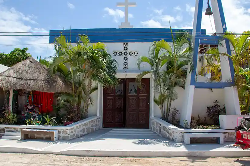 A church with a blue roof surrounded by palm trees, located in Holbox, Mexico, showcasing vibrant tropical scenery.