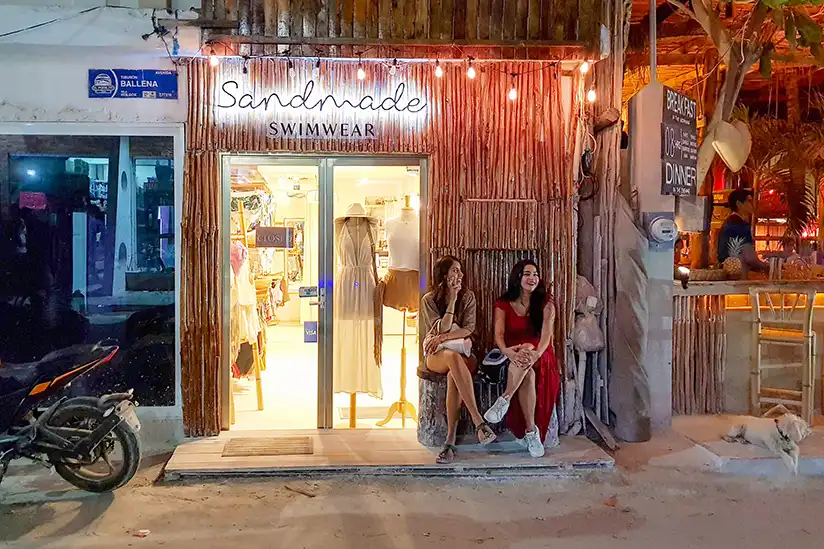 Two women sit relaxed at night in front of the Sandmade Swimwear boutique in Holbox.